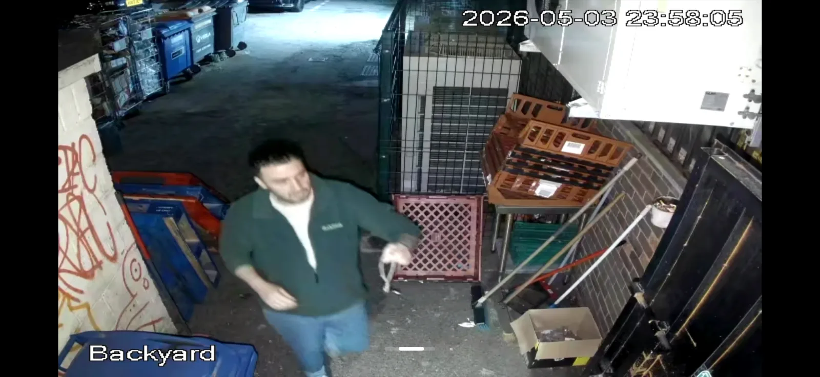Night-vision CCTV frame from a UK shop backyard, timestamp 2026-05-03 23:58, a person walking through the yard between stacked plastic crates near a metal cage.