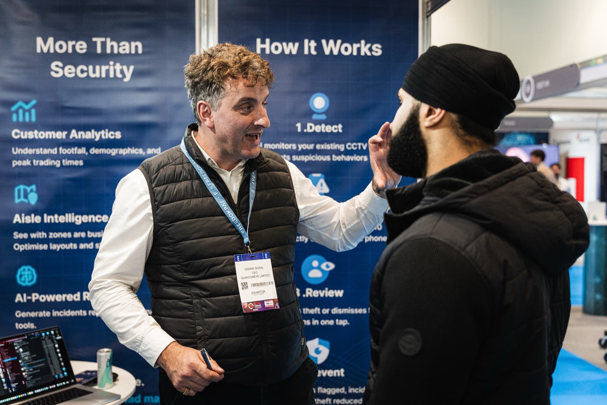 Osman Goral, QuantumEye CEO, in conversation with a visitor at a UK retail industry exhibition, in front of a branded backdrop reading 'More Than Security' and 'How It Works'.