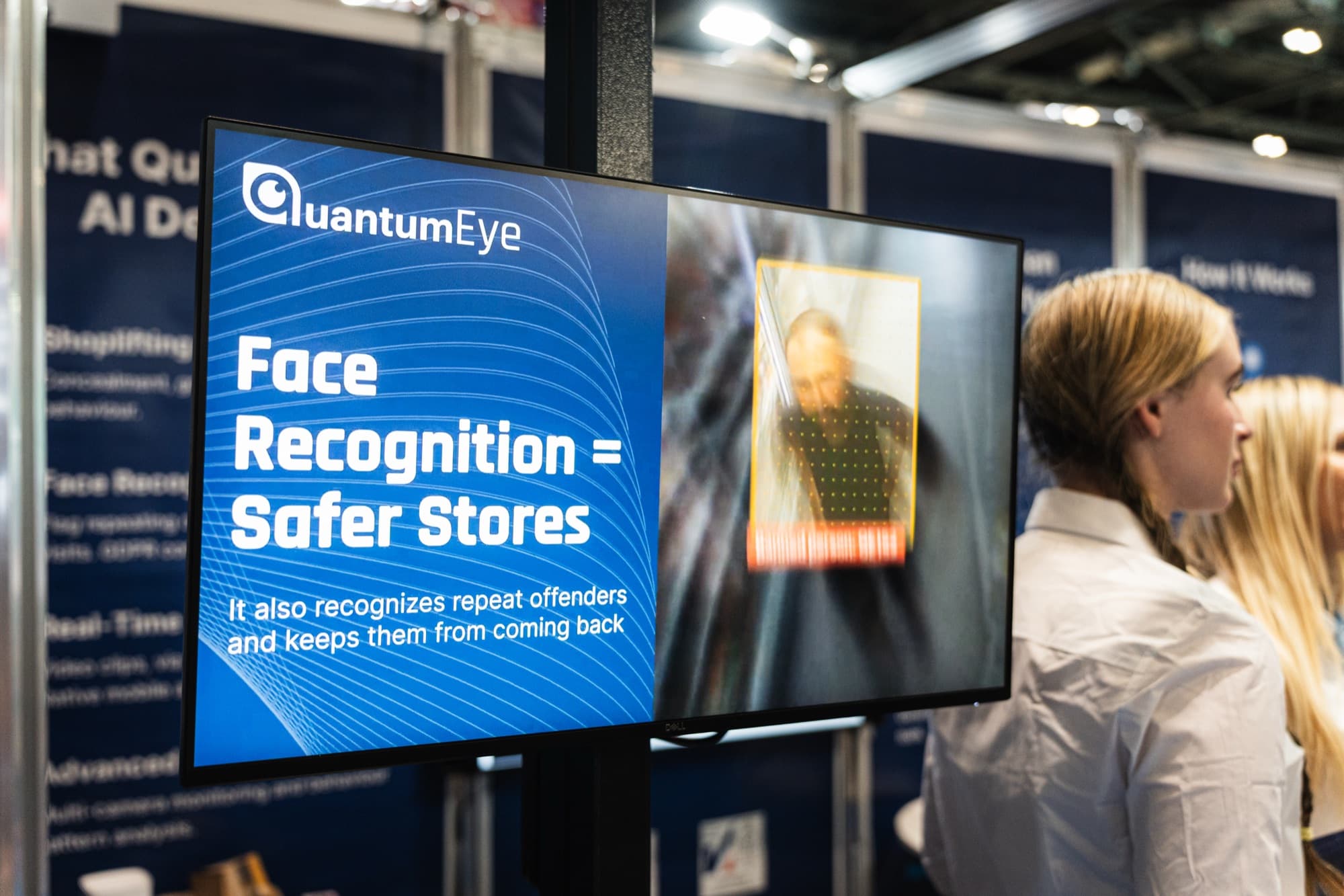 A QuantumEye exhibition-stand screen reading 'Face Recognition = Safer Stores, it also recognises repeat offenders and keeps them from coming back', shown to a visitor.