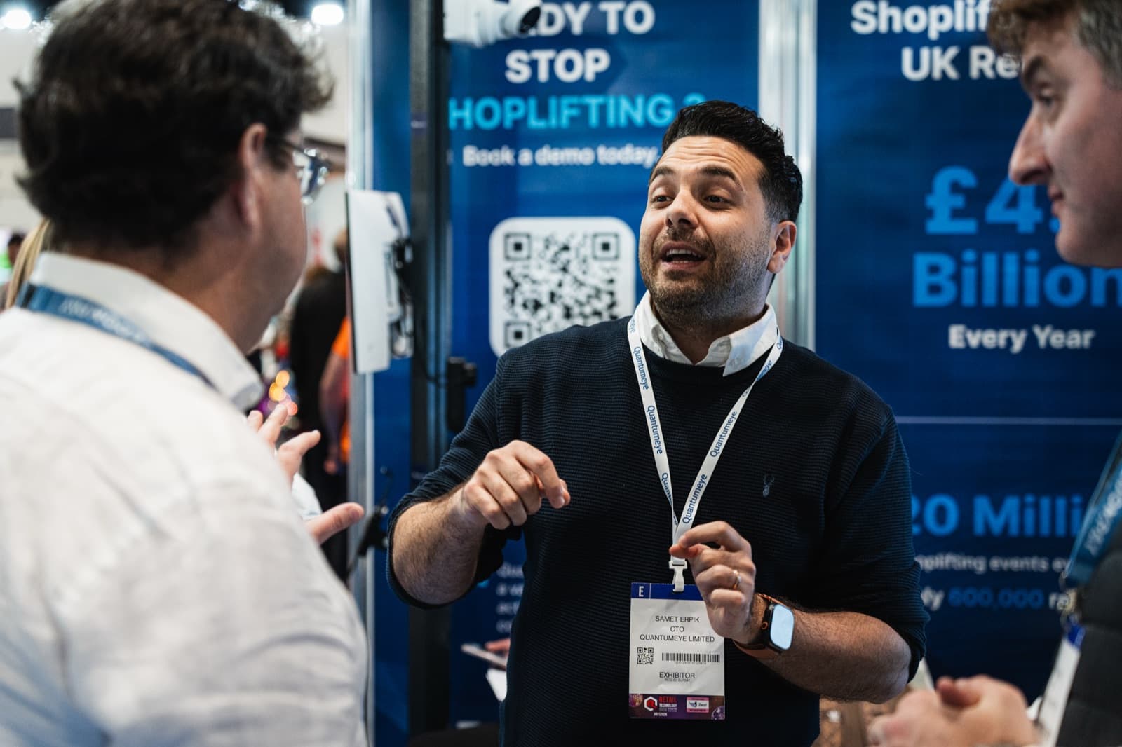 Sam Erpik, QuantumEye co-founder and CTO, talking with visitors at the stand, in front of a 'Ready to stop shoplifting?' backdrop citing the UK's £4bn / 20 million annual shoplifting figures.
